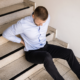 A man in pain sitting at the bottom of a stairwell holding his back after falling due to faulty construction