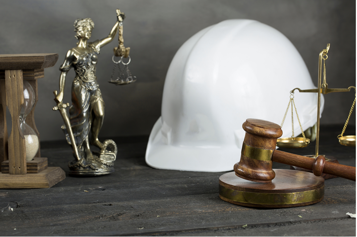 A construction hat sitting on a desk next to the scales of justice and a judge’s gavel representing Construction Law